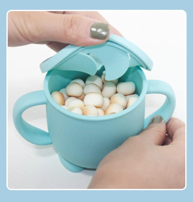 A hand is holding a light blue silicone snack cup with a lid, filled with small white balls for babies and toddlers.