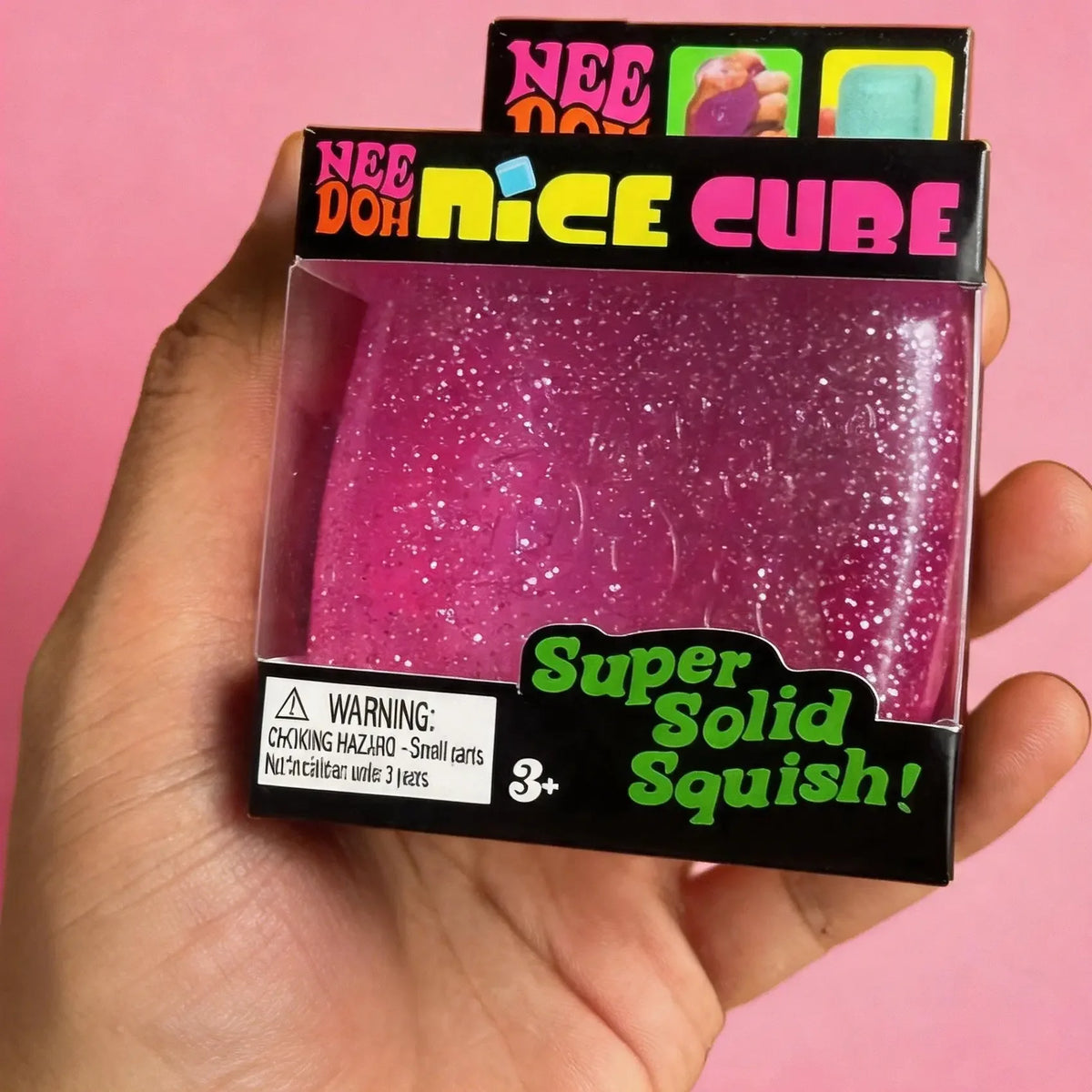A hand holds a NeeDoh Nice Cube sensory fidget toy which is a glitter ice cube in a black box.
