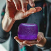 A hand holds a vibrant purple glitter ice cube sensory fidget toy against a dark background.