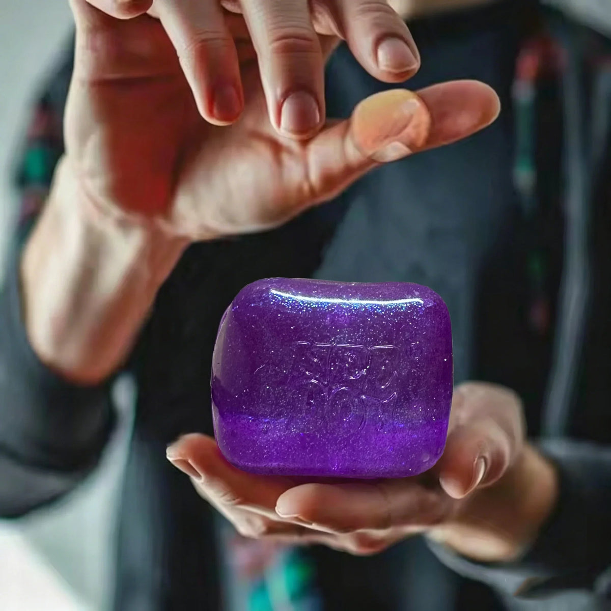 A hand holds a vibrant purple glitter ice cube sensory fidget toy against a dark background.