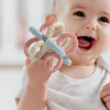 A baby is holding a silicone teether set with pink and white rings and a blue rattle toy.