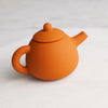 An orange silicone pretend play tea set teapot with a handle and spout sitting on a white surface.