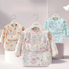 A light floral patterned toddler feeding bib with long sleeves and a pocket is displayed on a hanger.