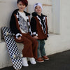 Two children wearing brown vests and checkered black and white outfits standing in front of a white wall.