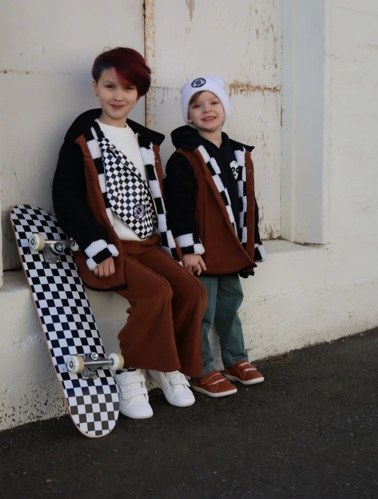 Two children wearing brown vests and checkered black and white outfits standing in front of a white wall.