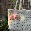 A handmade crochet mini pig keychain in pink and red is displayed on a light beige woven bag.