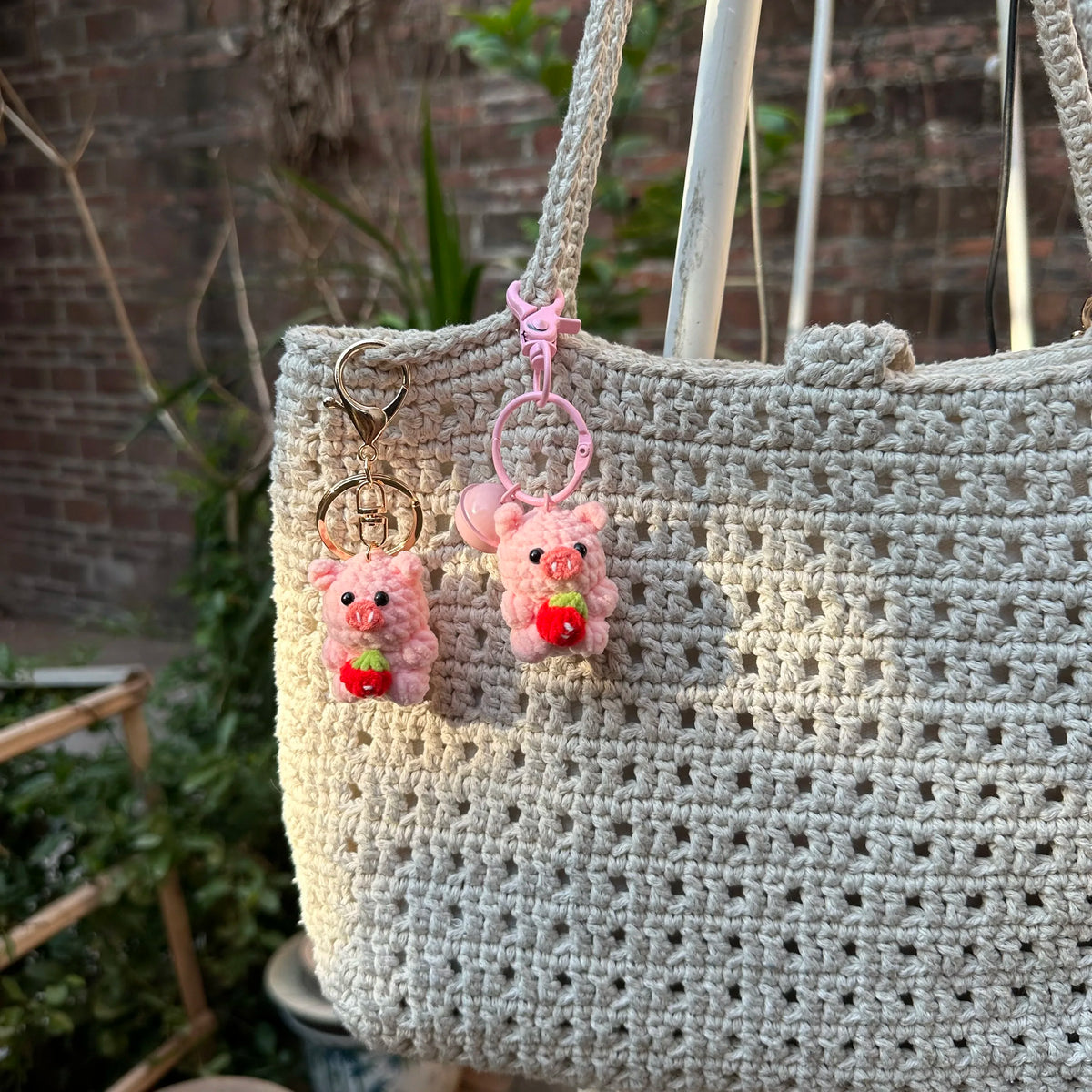 A handmade crochet mini pig keychain in pink and red is displayed on a light beige woven bag.