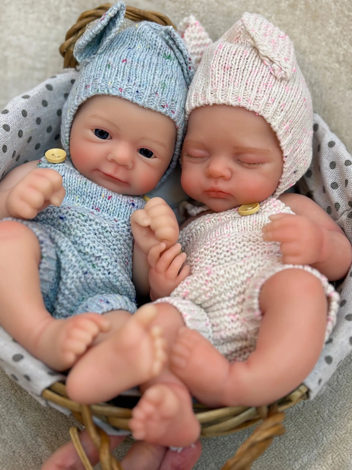 Two newborn silicone reborn dolls wearing knitted hats and outfits sitting in a woven basket