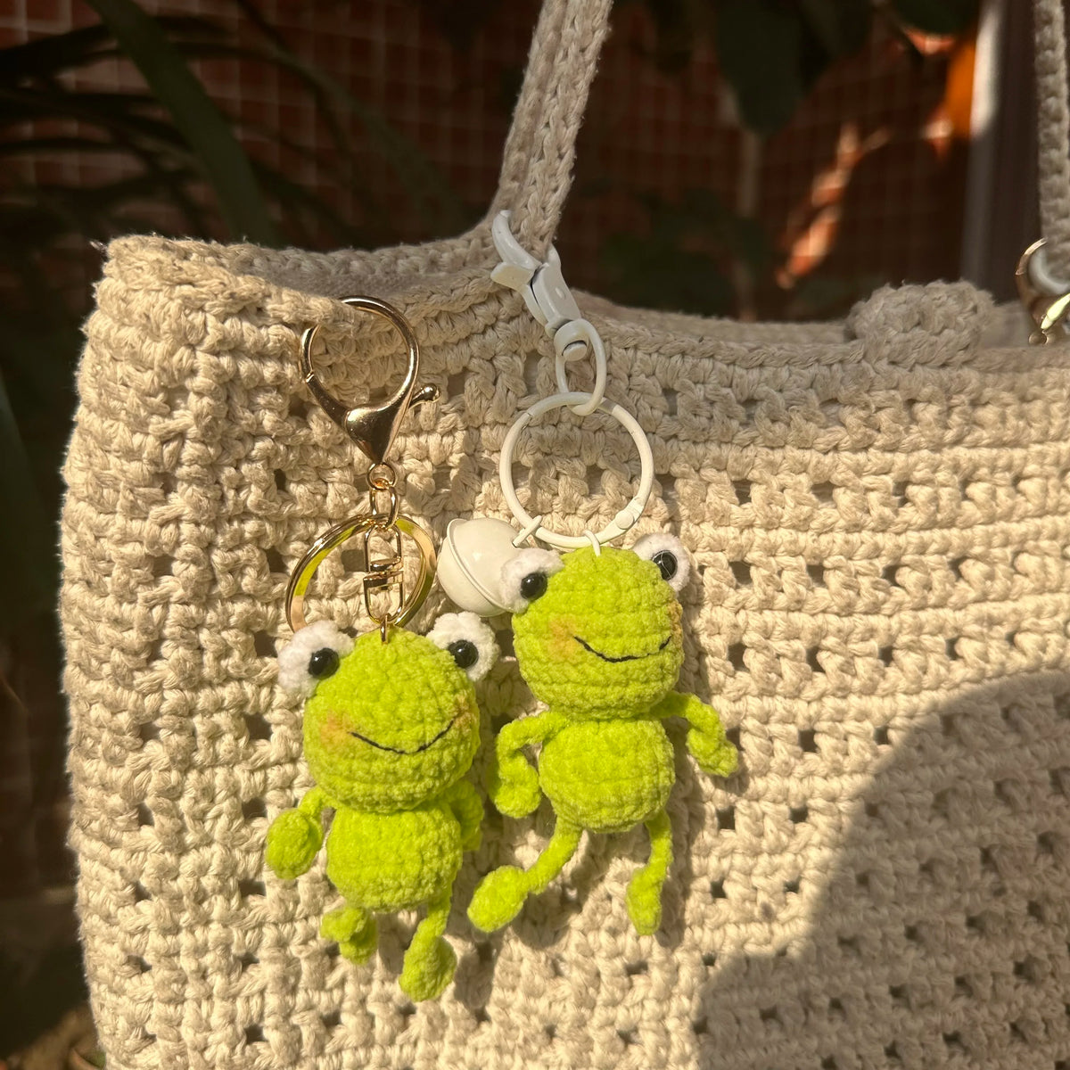 A handmade crochet frog keychain with yellow-green body and white accents hanging from a light beige knitted bag.