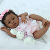 A newborn baby doll with dark skin, wearing a pink dress and a flower headband, lying on white fabric.