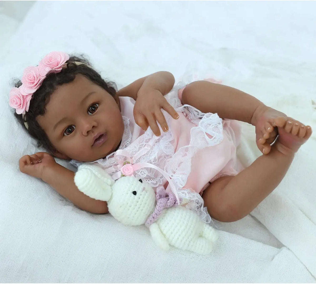 A newborn baby doll with dark skin, wearing a pink dress and a flower headband, lying on white fabric.