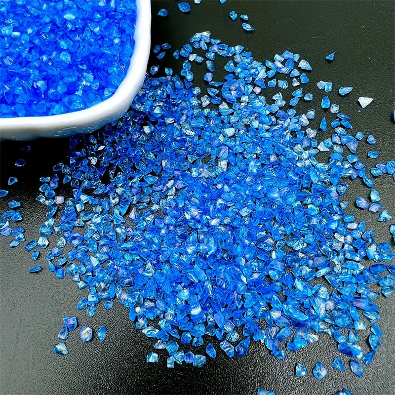 A pile of bright blue crushed glass crystals scattered on a dark surface next to a white bowl.