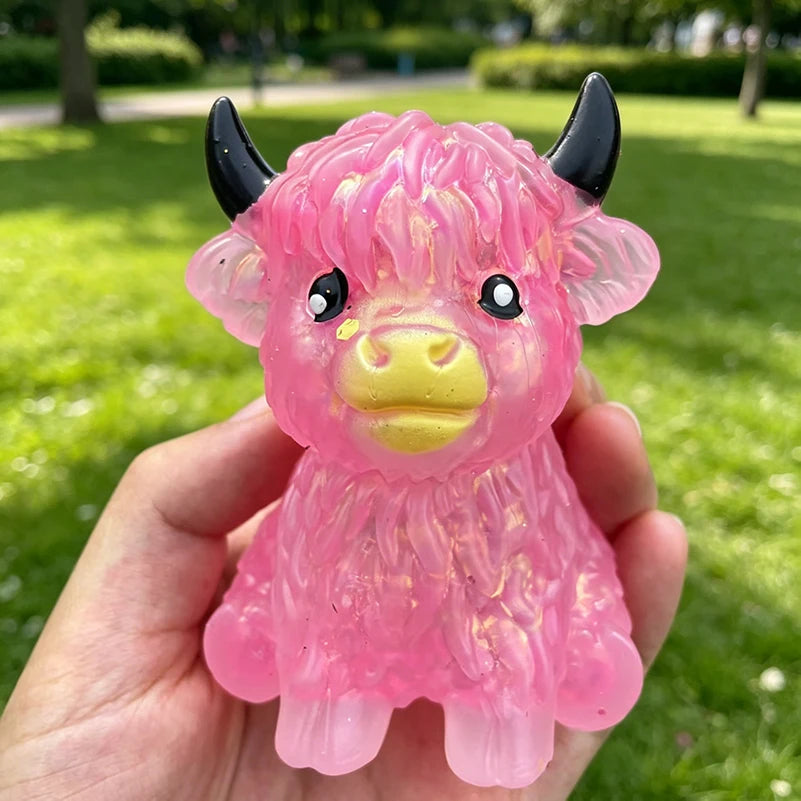 A hand holds a pink glitter highland cow squishy stress relief fidget toy with black horns.