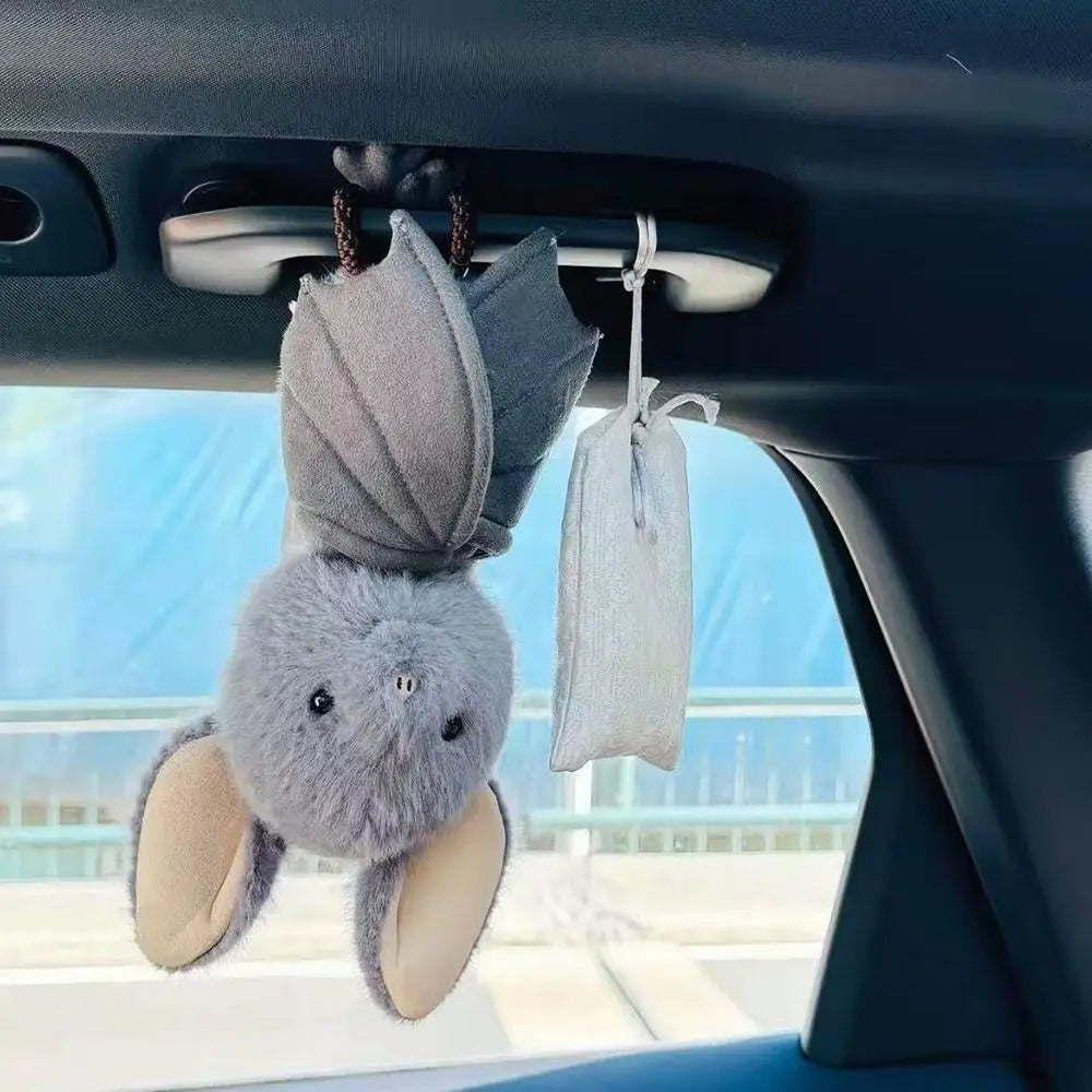 A grey bat plush toy is hanging from a car visor, with another light grey fabric pouch nearby.