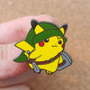 A yellow Pokémon enamel pin featuring a green hat and a brown belt, held by a person's finger.