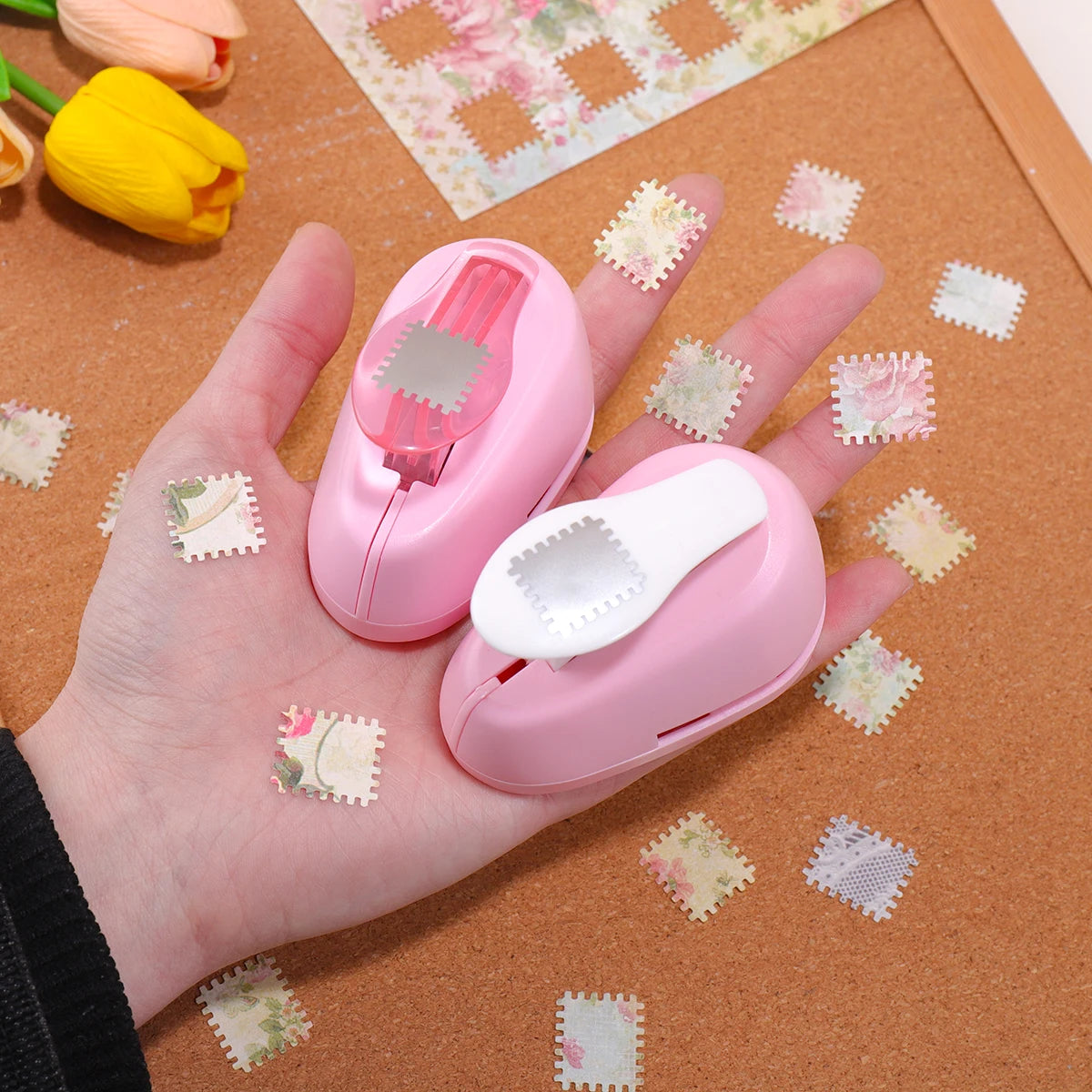 A hand holds a pink lever action embosser tool with a white cap, surrounded by small pink and white floral stickers on brown 