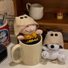 A beige plush Snoopy character is seated in a mug, with other small plush items visible on a wooden shelf.