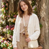 A young woman is holding a cream-colored knitted cardigan over a beige top, with a handmade crochet sloth and squirrel plush 