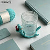 A handwoven turquoise crochet lizard coaster sits next to a glass and books on a white surface.