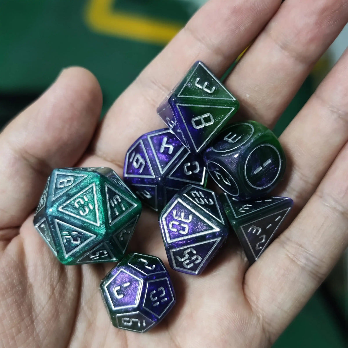 A hand holds a set of seven resin polyhedral dice with a display frame on a green surface.