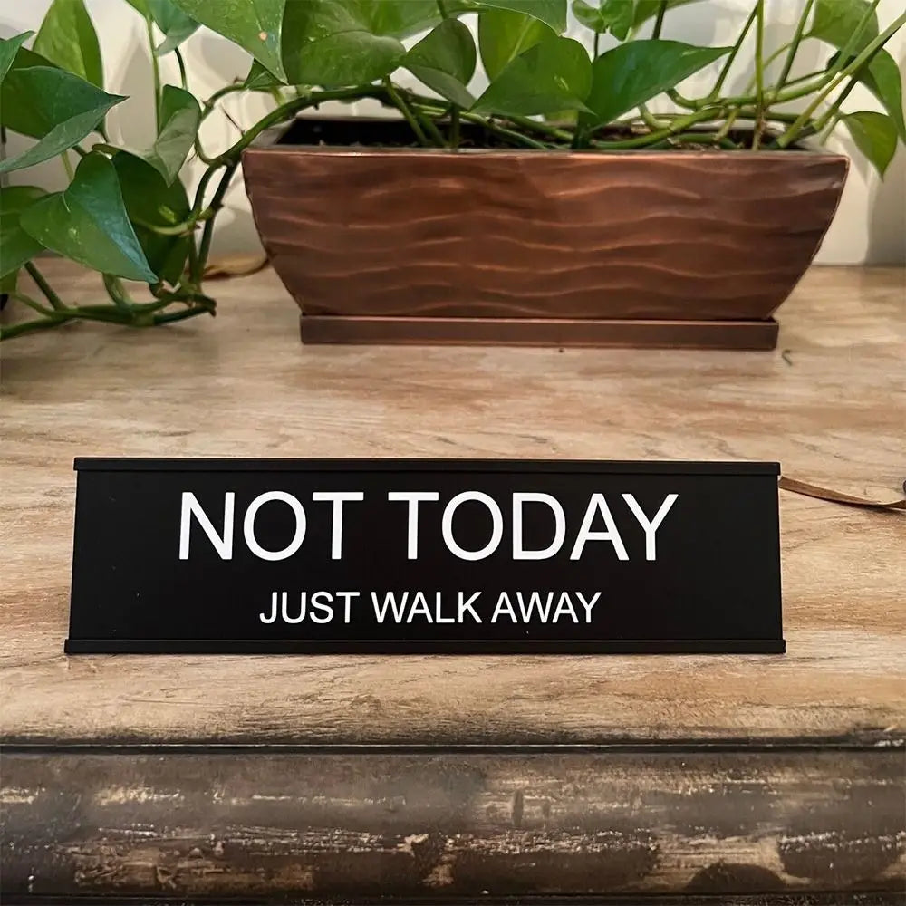 A black desk sign with white text that reads NOT TODAY JUST WALK AWAY placed in front of a potted green plant.