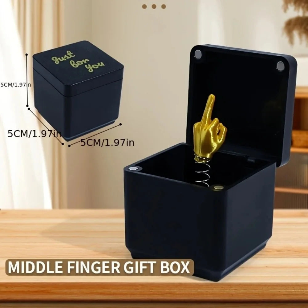 A black pop-up gift box with a gold middle finger figurine is shown on a wooden surface.