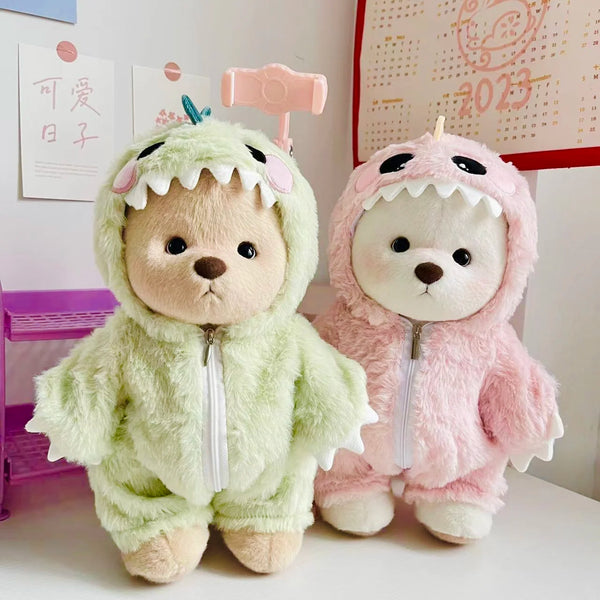 A pair of fluffy bear dolls dressed in dinosaur jumpsuits, one green and one pink, standing on a white surface.