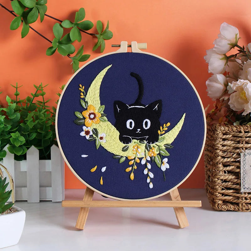 A dark blue embroidery featuring a crescent moon and a black cat silhouette with yellow and white floral accents.