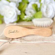 A light wooden baby brush and comb set with an engraved name resting on a beige cloth.
