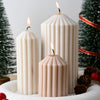 A grouping of three white candles in varying heights with lit flames on a white surface.
