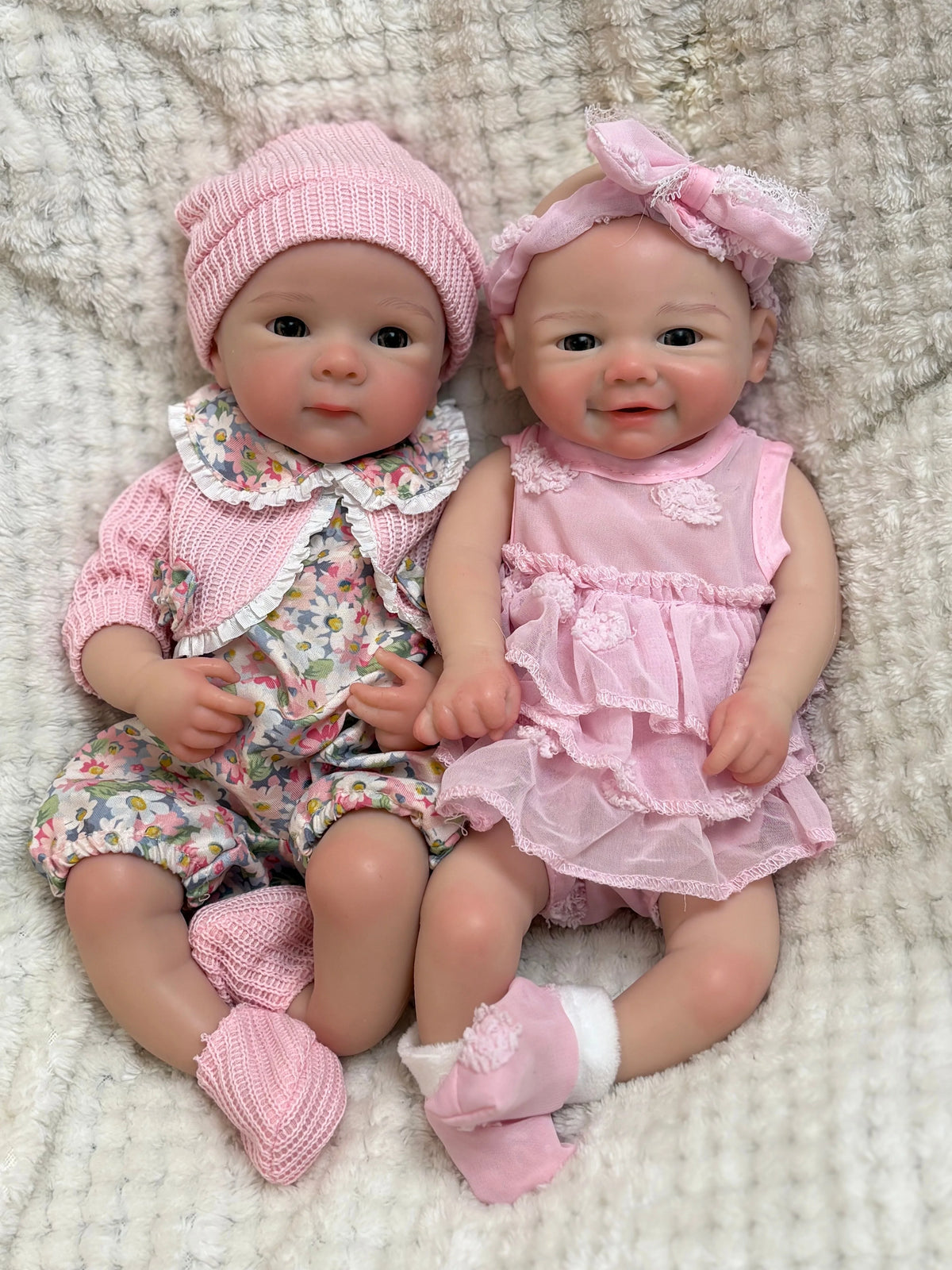 A realistic soft silicone reborn baby doll with a pink outfit and bonnet sitting on a light textured surface.