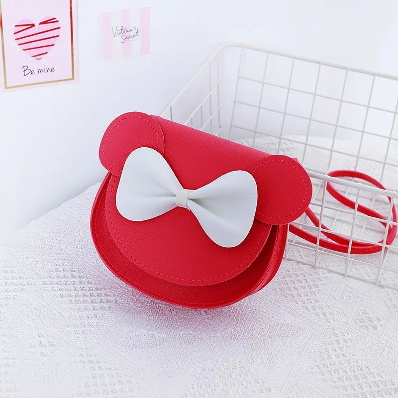 A red kids' hard shell crossbody bag with white bow and mouse ears resting on a white mesh basket.