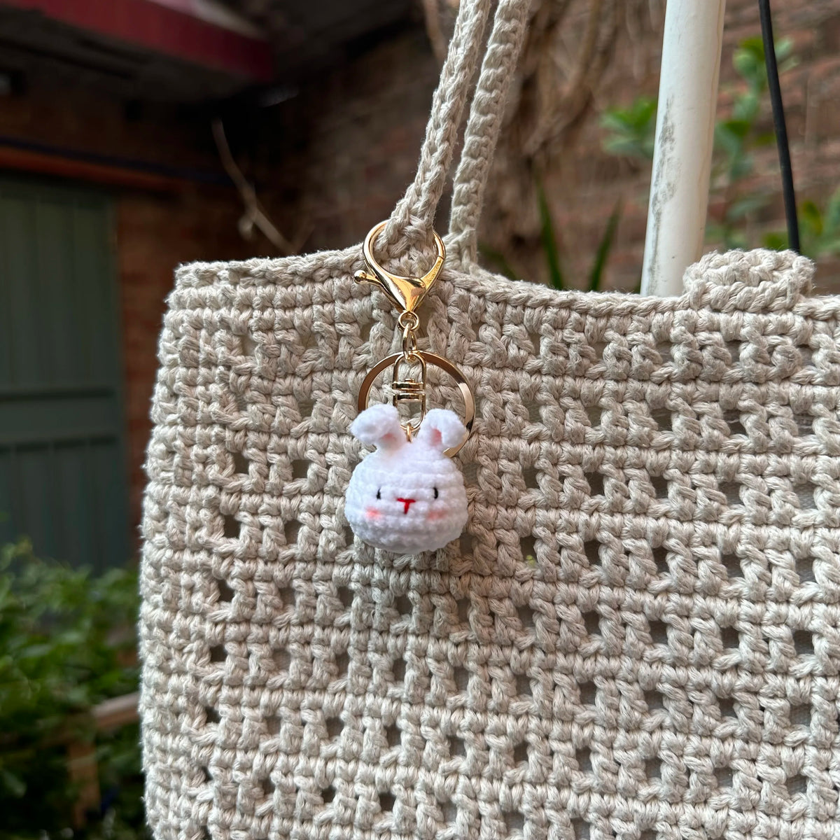 A light beige crocheted handbag with a gold-toned keychain featuring a small white rabbit charm.