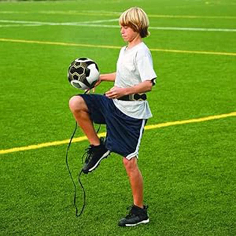 A young male soccer player wearing a white shirt and dark shorts is practicing with an adjustable solo kick training belt on 