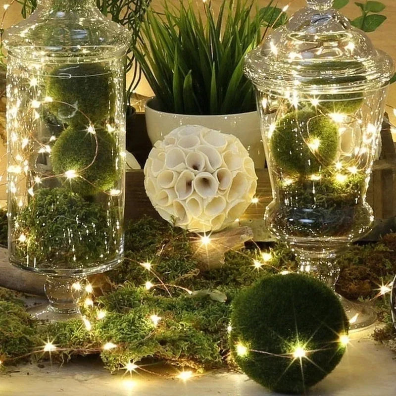 Battery-powered fairy lights illuminating moss balls and greenery in a glass jar