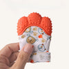 A hand holds an orange silicone teething mitten with a white fabric interior featuring cute animal patterns.