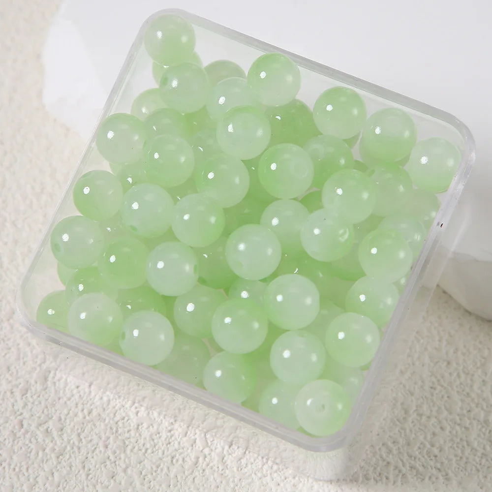 A clear plastic container filled with small, round, light green imitation jade glass beads for jewelry making.