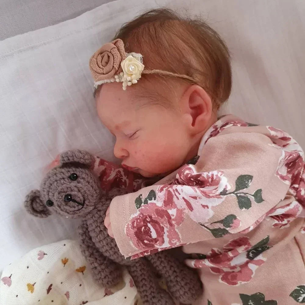 A newborn baby with light brown hair is sleeping, wearing a pink floral outfit and a rose headband, holding a small knitted e