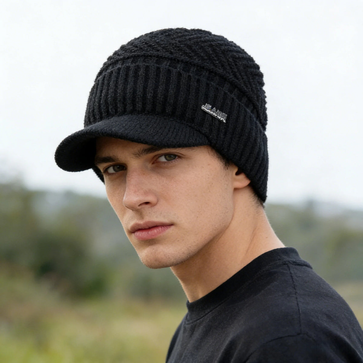 A male model wearing a black knit beanie with a brim and a black shirt against a blurred outdoor background.