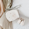 A cream quilted diaper bag with pink embroidery rests on a white surface next to a pair of matching slip-on shoes.
