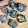 A collection of handcrafted resin polyhedral dice in shades of blue and teal with gold numbers resting on lined paper.