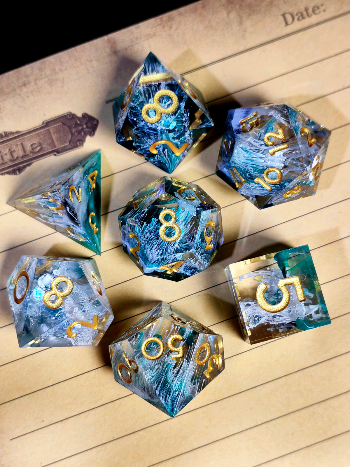 A collection of handcrafted resin polyhedral dice in shades of blue and teal with gold numbers resting on lined paper.