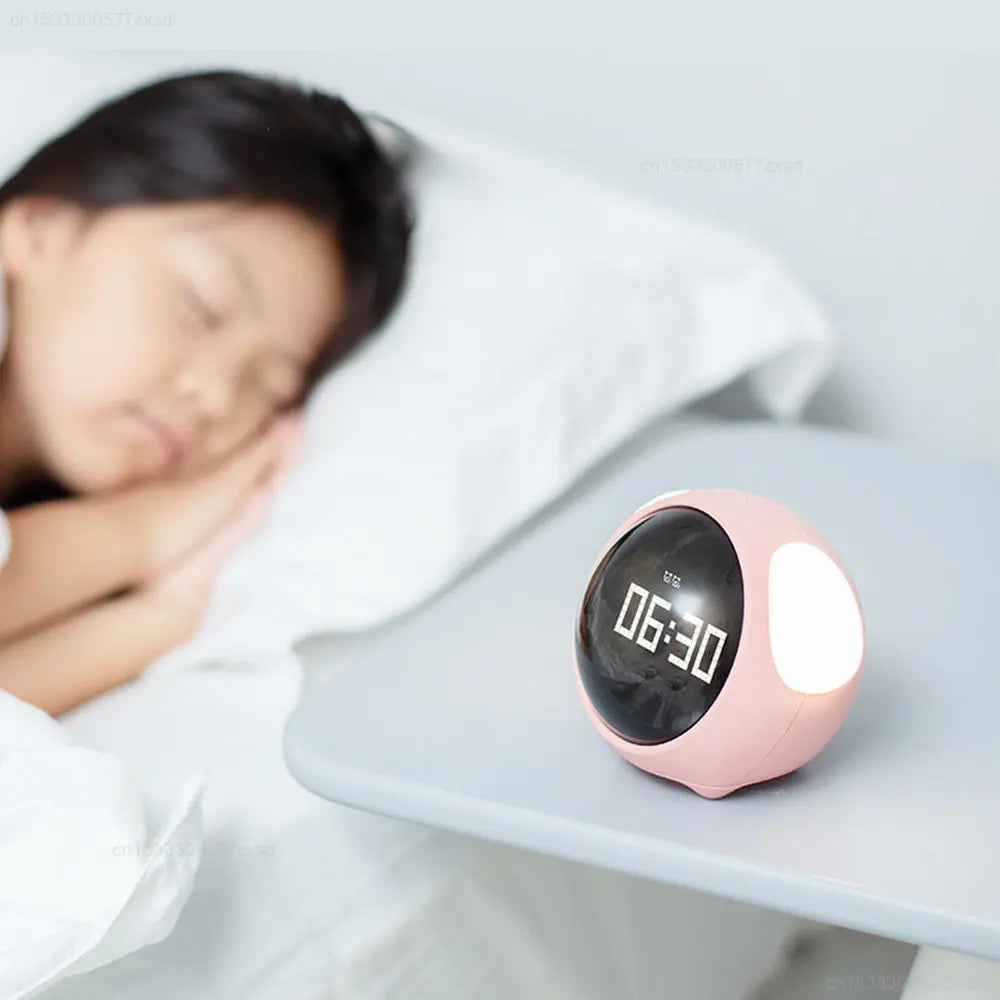 A pink emoji alarm clock with a digital display showing 06:30 is resting on a white surface next to a person sleeping.