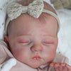 A newborn baby doll with a light beige bow headband sleeping in a pink knitted outfit.