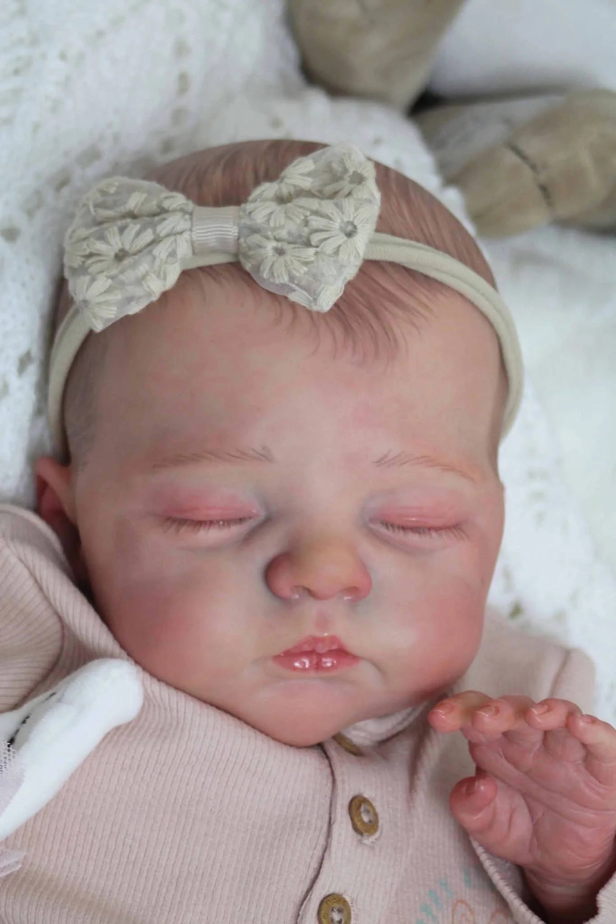 A newborn baby doll with a light beige bow headband sleeping in a pink knitted outfit.