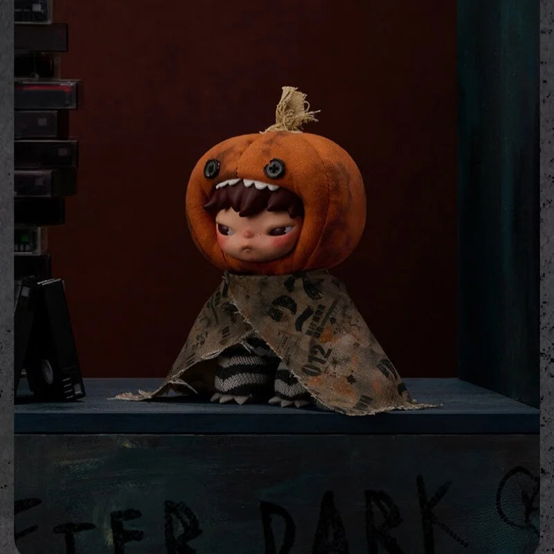 An orange plush doll with a stitched mouth and hair, wearing a patterned fabric drape, displayed on a dark surface.