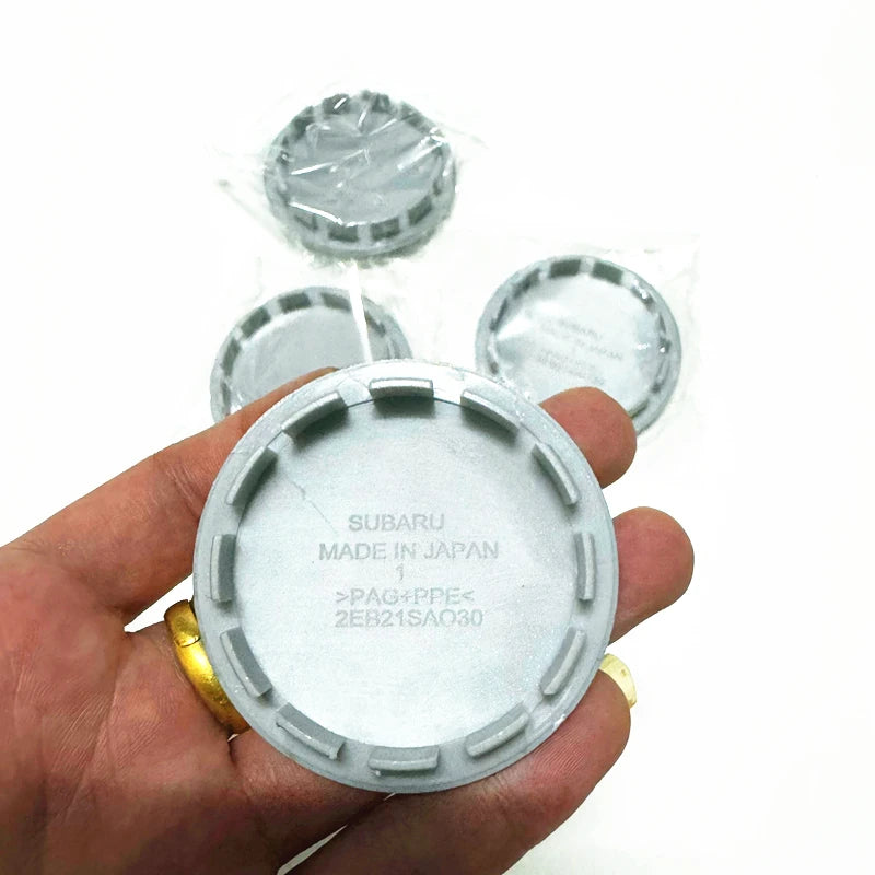 A hand holds a set of four silver car wheel hub center caps for Subaru models.