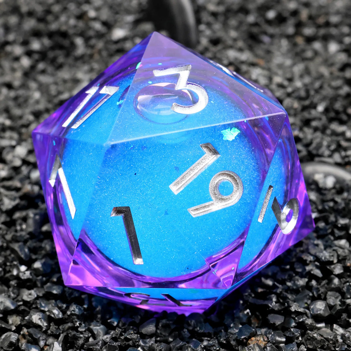 A translucent purple and blue resin D20 die with a large liquid swirl design resting on black gravel.
