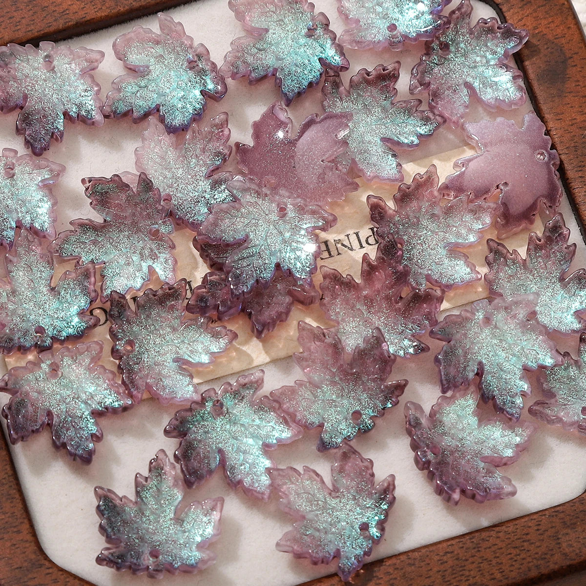 Pink and teal maple leaf glass beads arranged on a wooden surface for DIY jewelry making