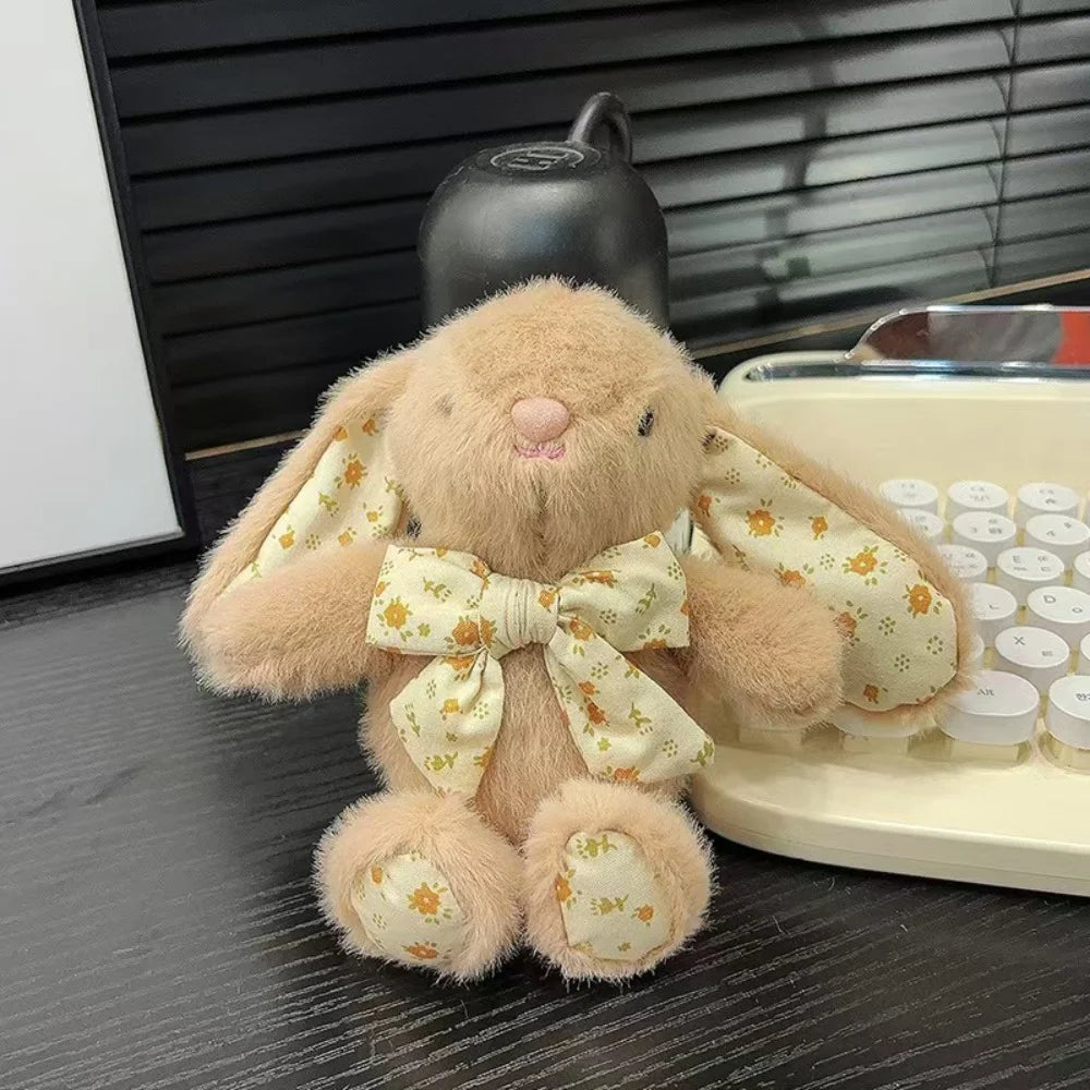 A soft, light brown bunny plush toy with a yellow floral bow charm sitting on a dark surface.